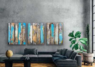 Detailfotografie der Oberfläche eines Holztisches in blau-braun-gelben Farbtönen als drei großformatige Leinwandbilder an grauer Wand über dunkelgrauer Couch.
